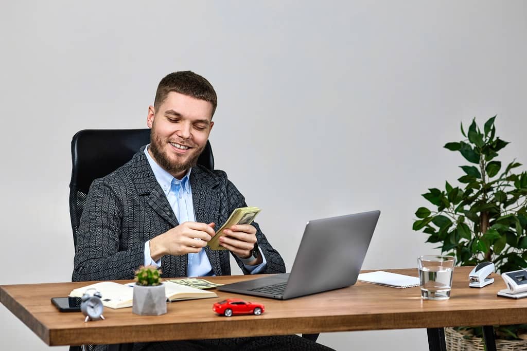 Article campagne social ads - Un homme souriant,  assis à son bureau devant son ordinateur et tenant une liasse de billets 