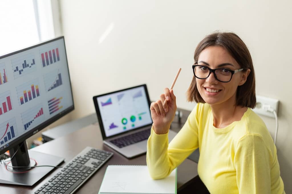 Article gestionnaire de publicité - Une femme souriante à la caméra, assise à son bureau devant un ordinateur affichant des graphiques