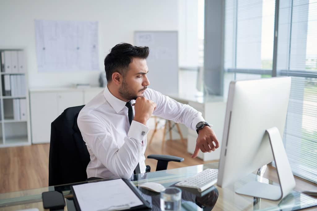 Article social ads expert - Un homme regardant l'heure sur sa montre, assis à son bureau, devant son ordinateur
