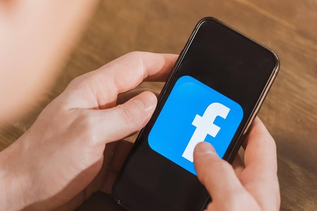 Article social ads prix - Vue en plongée sur les mains d'une personne tenant un téléphone affichant le logo de Facebook