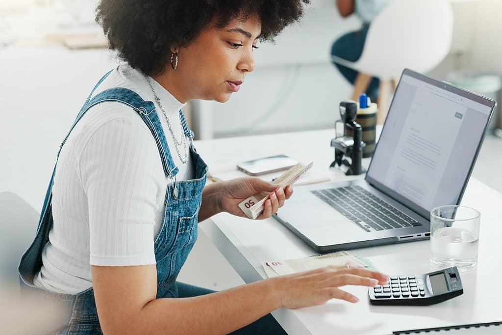 Article social ads prix - Une femme assise à son bureau en train d'utiliser une calculatrice avec sa main droite et tenant des billets dans sa main gauche